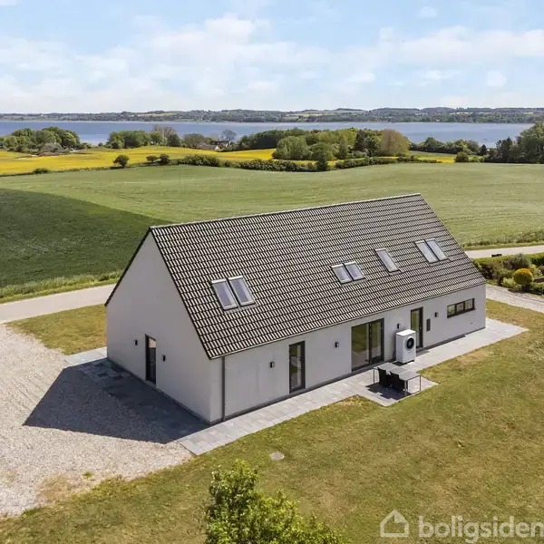 En hvid, moderne etplansbygning med skråt tag står stille på en velplejet græsplæne. Den er omgivet af åbne marker, og der er en fjord i baggrunden under en blå himmel.