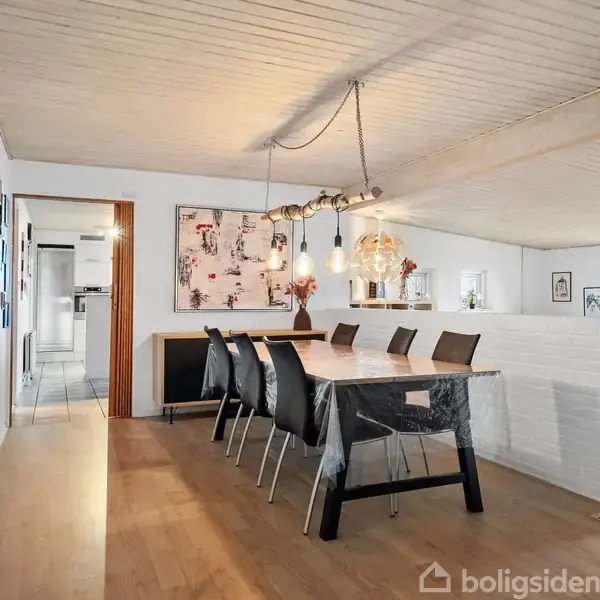 Spisebord med otte sorte stole står i en lys spisestue med trægulv og hvidt loft. Lamper hænger over bordet, væggene er dekoreret med billeder, og en åbning fører til køkkenet.