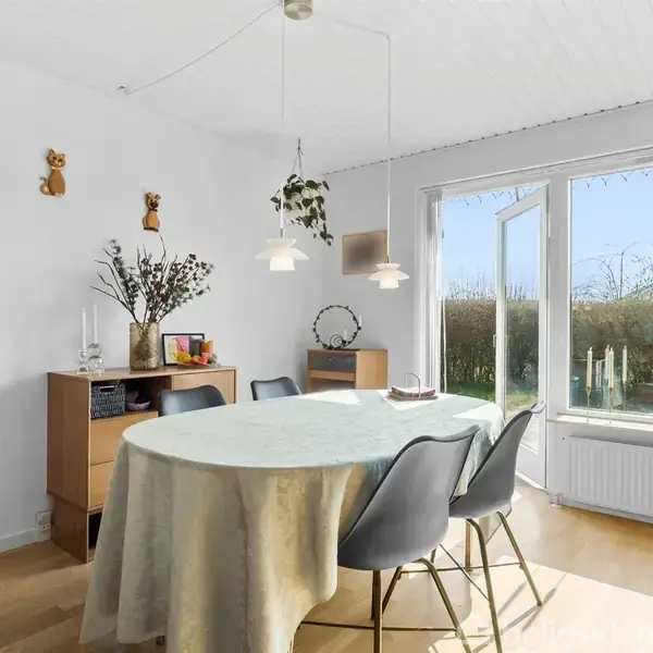 Ovalt bord dækket med dug står i en lys spisestue. Omkring bordet er der stole, og i baggrunden en åben dør til haven. En kommode med pynt ses til venstre.