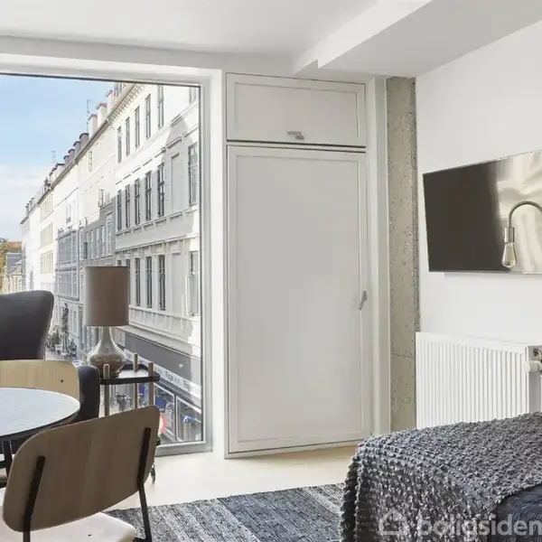 Et moderne værelse med en rund spisebord med stole, en lænestol ved siden af et stort vindue med gadeudsigt, en seng i forgrunden, og en vægmonteret fladskærm.