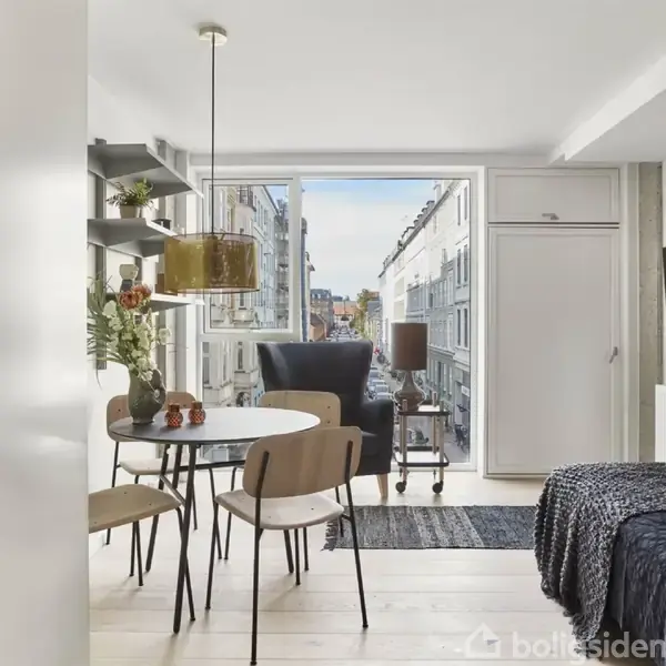 Et moderne, lyst rum indeholder en rund spisebord med stole. Ved siden ses en seng og en mørk lænestol. Et stort vindue viser udsigt til byens gader.