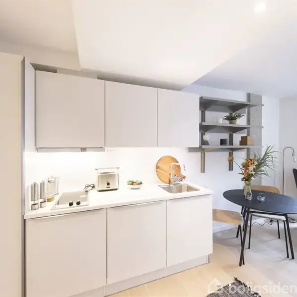 Køkken med hvide skabe står ved siden af en hvid vask. Over bordpladen er en hylde med dekorative genstande. En rund sort bord med blomster ses ved siden af en blå stol.