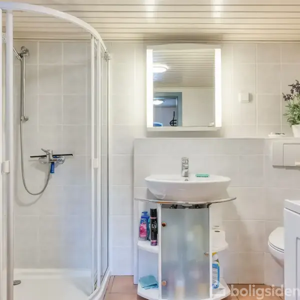 En moderne badeværelsesindretning med en brusekabine, vask med spejl og hylder med artikler. Omgivet af lyse fliser, en vaskemaskine og en plante ved toilettet.