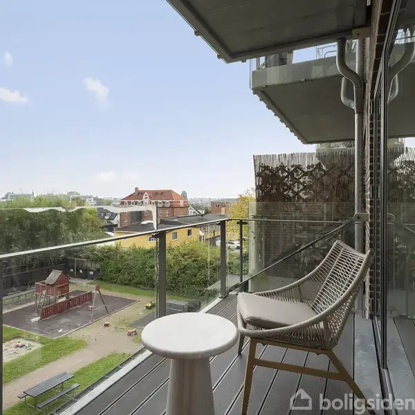 En balkon med en stol og et lille bord vender ud mod en legeplads og grønne områder. Bygninger er synlige i baggrunden under en klar, blå himmel.