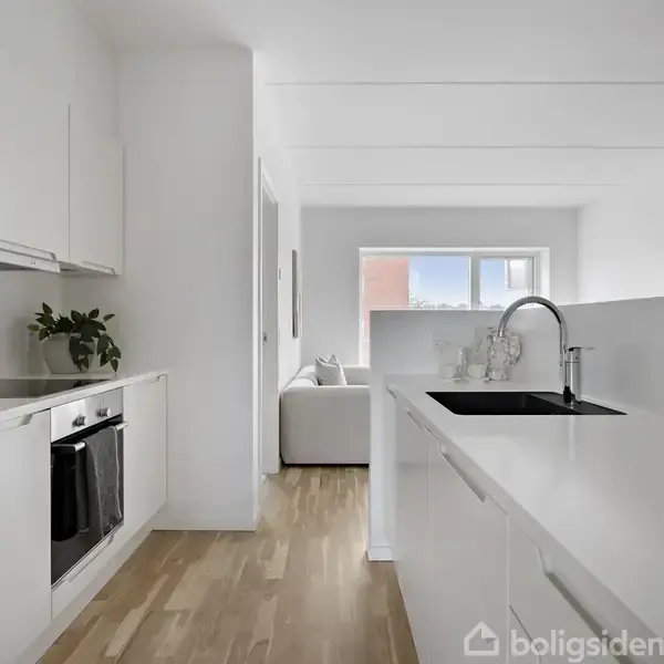 Et hvidt, moderne køkken med indbyggede skabe og en vask i en ø, fører ind til en stue med lysindfald fra et vindue. Trægulve dækker hele området.