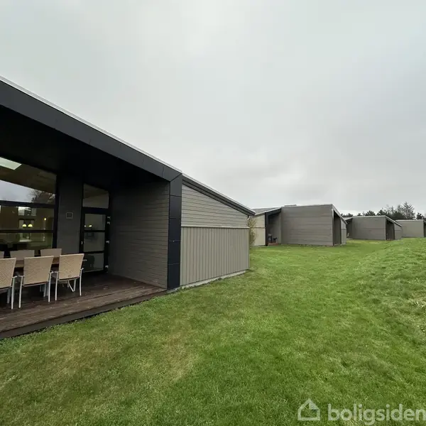 Et rækkehuskompleks med moderne flade tage står på en grøn græsplæne. Der er udendørs spisebordsstole ved en terrasse. Baggrunden viser flere ens huse og træer.