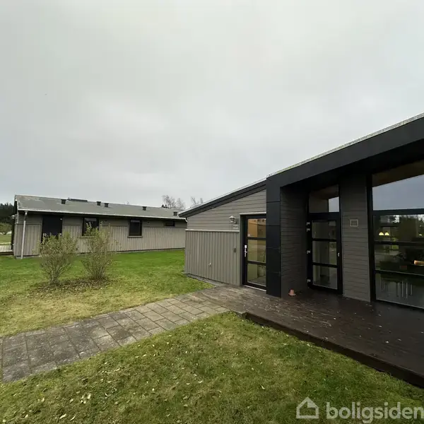 Bygningerne har flade tage og store vinduer, der reflekterer det overskyede himmel. Foran ses en flisebelagt gangsti og grønne græsplæner i et boligområde.