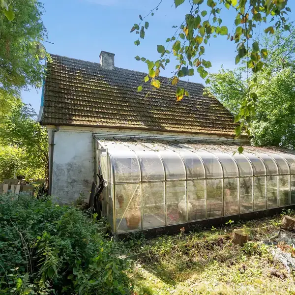 Et gammelt hus med en tilstødende glaskonservatorium står stille i en frodig have. Træer og buske omkranser det, mens solen skinner klart på taget.