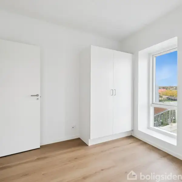 Et hvidt garderobeskab står i et rum med trægulv ved siden af et stort vindue. En åben dør fører til et tilstødende rum med dagslys udenfor.