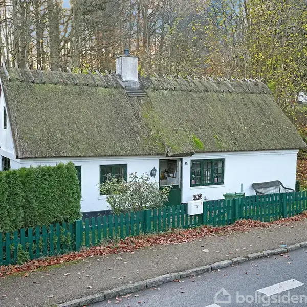 Et lille hvidt hus med stråtag står stille, omgivet af en grøn hæk og hegn. Baggrunden består af efterårsfarvede træer og en rolig vej foran huset.