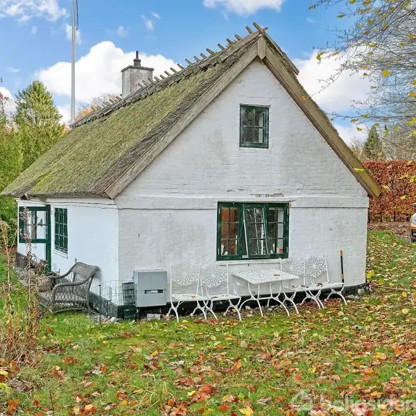 Huset er et hvidt, gammelt bindingsværkshus med stråtag, omgivet af grøn græsplæne og faldende efterårsblade. En gul bil er parkeret i baggrunden ved siden af træer.