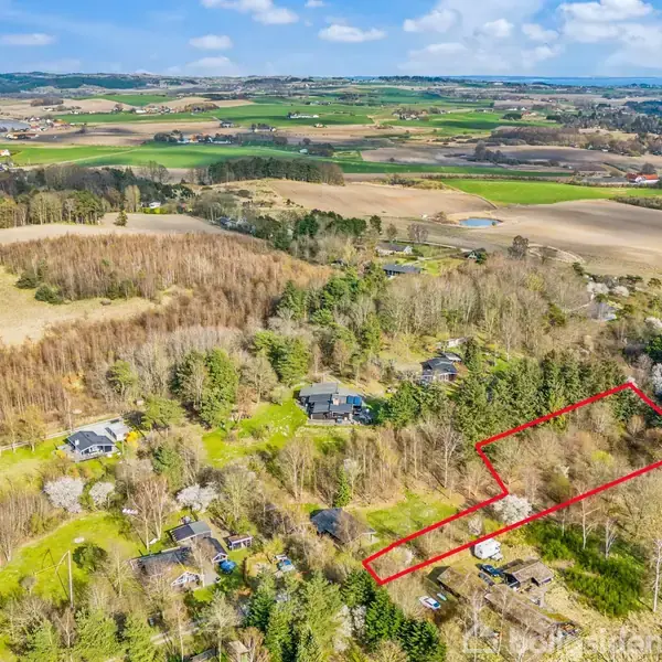 En rektangulær grund omgivet af skov og spredte huse, markeret med en rød streg. I baggrunden ses åbne marker og en kystlinje.