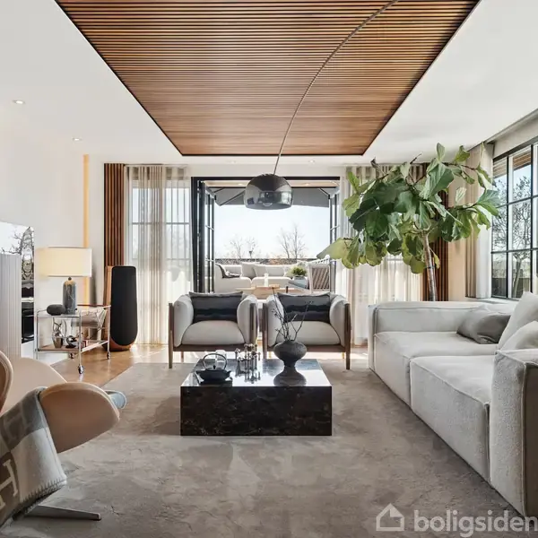 Elegant stue med moderne møbler, hvor en sofa og stole omkranser et sofabord. Store vinduer giver lys, mens planter og lamper bidrager til et stilfuldt og hyggeligt miljø.
