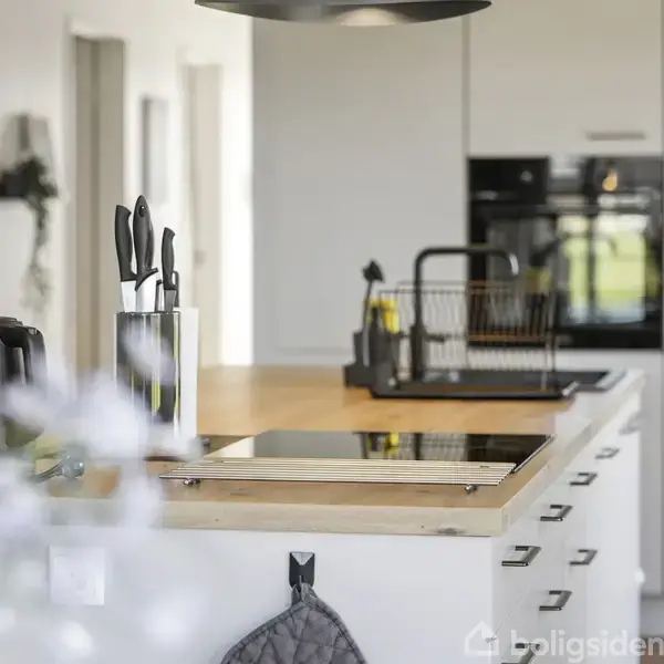 Køkken med komfur på træbordplade, knivblok og elkedel. Baggrunden viser opvaskestativ og indbyggede skabe i et lyst rum med blomster i forgrunden.