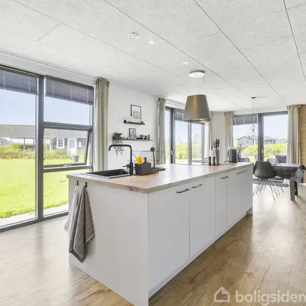 Et moderne køkken med en hvid køkkenø, der står centralt i et lyst, åbent rum med store vinduespartier, der giver udsigt til en grøn have. Der er en stor lampe over øen, og rummet åbner op til en stue med sofa og TV.