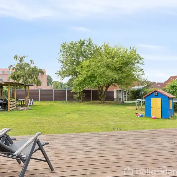 To liggestole står på en træterrasse ved siden af en grøn græsplæne. I baggrunden er et farverigt legehus, et gyngestativ, træer og en havepavillon.