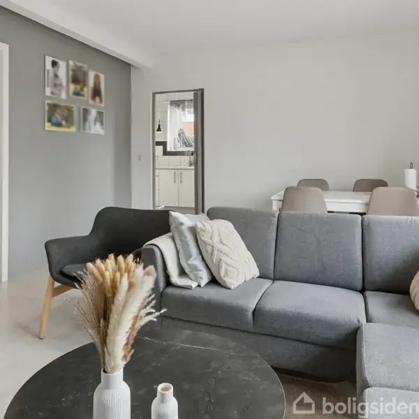 Gråt sofasæt er centralt i en lys stue, omgivet af et rundt sofabord. Til venstre hænger billeder på en væg, og en dør fører til et farverigt værelse. En spiseplads med fire stole ses bagved.