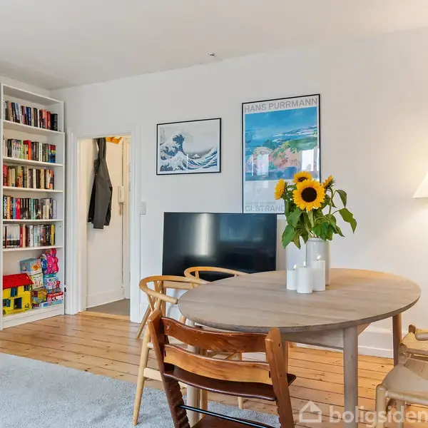 Et rundt bord med solsikker i vase står i et lyst stuehjørne. Ved siden af er en reol fyldt med bøger. En lampe er tændt i hjørnet, og en fjernsynsskærm ses bagved.