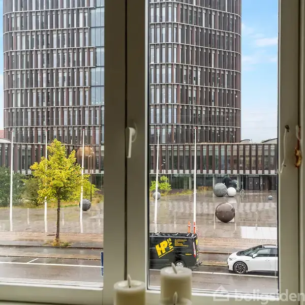 Et vindue viser en moderne, høj bygning med glasfacade udenfor. På pladsen foran står runde skulpturer, træer langs vejen, og biler er parkeret på gaden.