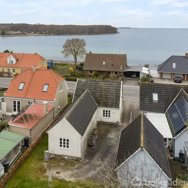 Flere huse med røde og mørke tage står på række ved vandet. En vej adskiller husene fra kysten. Grønne haver omringer bygningerne, og skoven ses i baggrunden.