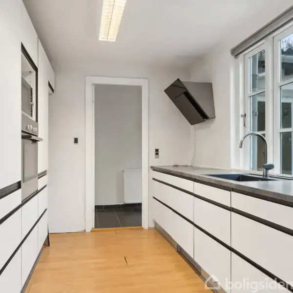 Køkken med hvide skabe og indbygget ovn, der står stille. Rummet har lyst trægulv og store vinduer, der slipper naturligt lys ind. En dør i baggrunden.