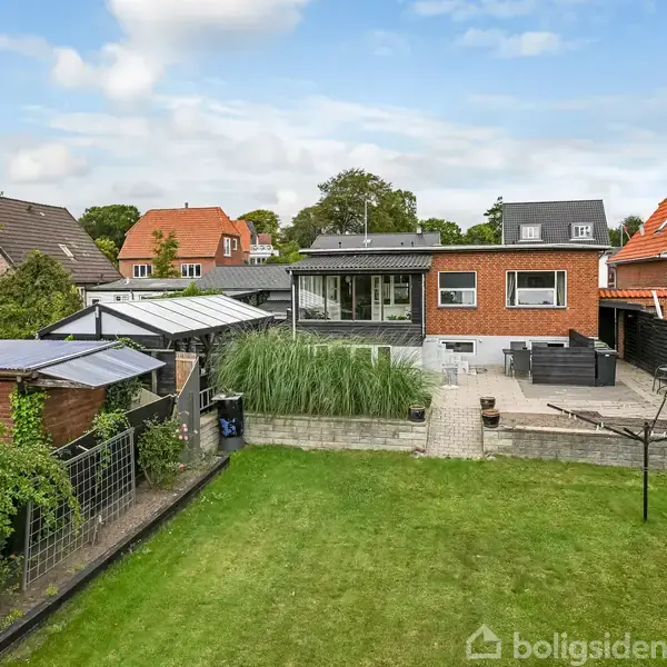 En rød murstensbygning står i et parcelhuskvarter. Foran er en stor græsplæne, terrasse og grønne planter. Flere huse og træer kan ses i baggrunden.