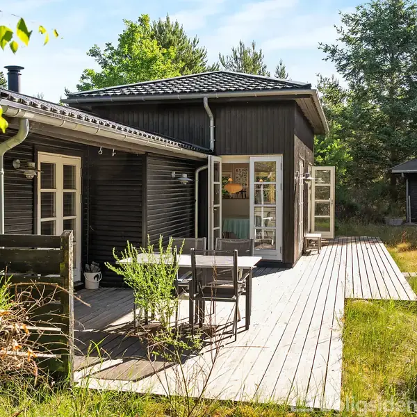 Sortmalet træhus med åbne døre og tilhørende terrasse med havemøbler. Terrasse omgivet af græs og træer, hvilket skaber en fredelig skovhave-atmosfære.