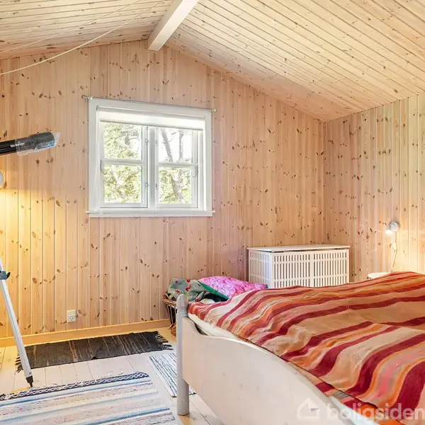 Et soveværelse med en teleskop på stativ ved vinduet. Sengen har en stribet dyne. Væggene er af lyst træ. En lampe og et kort hænger på væggen.