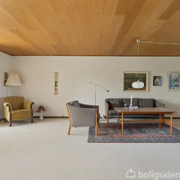 Stue med en brun sofa ved et træbord, flankeret af to lænestole. Gulvtæppe under bordet. Væglamper hænger ved hvide vægge med malerier. Store vinduer til venstre.