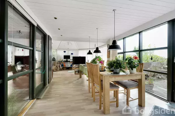 Spisebord med træstole og planter står i en lys stue med store vinduer, der giver udsigt til en terrasse. Loftslamper hænger over bordet, og et fjernsyn er i baggrunden.