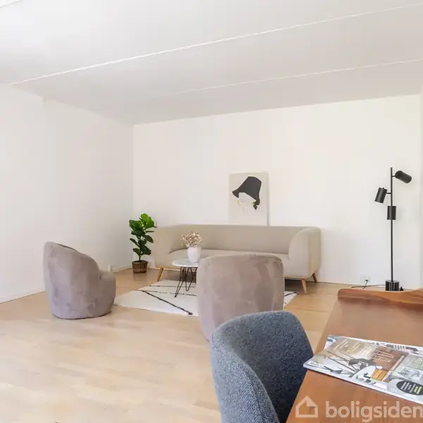 En moderne dagligstue indeholder en sofa, to stole og et bord, omgivet af lyse vægge. Et skrivebord med en stol er i forgrunden. En plante og en lampe tilføjer dekoration.