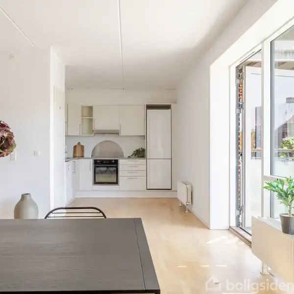 En moderne køkken-spisestue med hvide skabe og en ovn i baggrunden. Et sort bord med en vase med blomster står i forgrunden, og der er udgang til altan til højre.