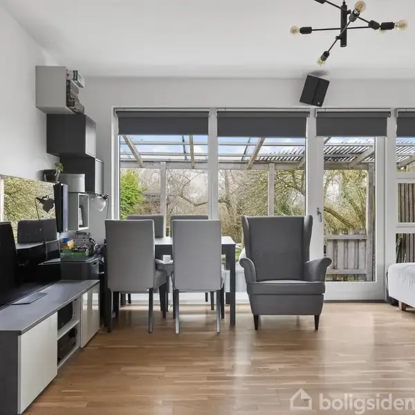 Stue med spisebord og stole, lænestol, sofabord og computerudstyr. Stuen har store vinduer med udsigt til en terrasse og have. Loftet har en moderne lysekrone.