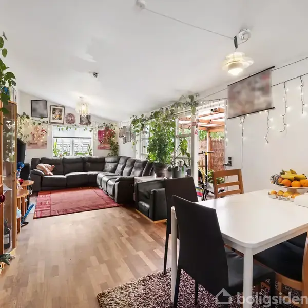 Stue med sorte lædersofaer i hjørnet, omgivet af grønne planter og dekorerede vægge. Et spisebord med stole står ved siden af. En frugtskål ses på bordet.