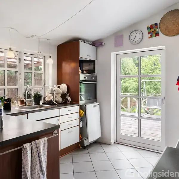 Køkken med mørke træskabe og hvide overflader. Køkkenet er rodet med opvask på bordet. Store vinduer og en glasdør åbner ud til en terrasse med grønt udsigt.
