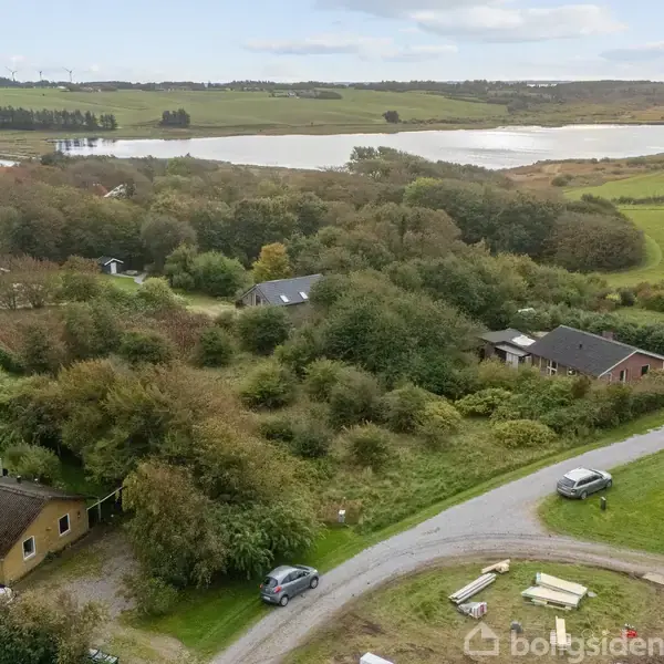 En landsby med huse blandt træer og buske. Veje og biler er synlige i forgrunden. En sø og marker er i baggrunden under en klar himmel.