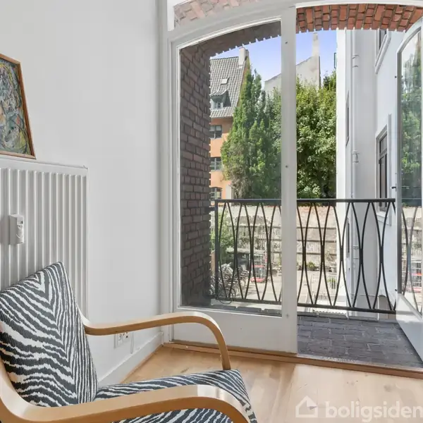 Stol med zebrastribet betræk står ved siden af en radiator med kunst ovenpå. Åbent vindue fører til en balkon med udsigt til træer og bygninger udenfor.