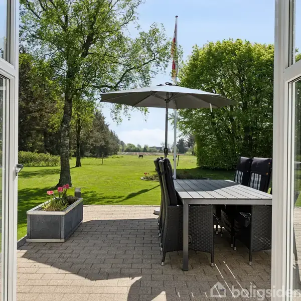Et bord med stole står under en parasol på en terrasse. Terrassen åbner ud mod en frodig have med grøn plæne og træer i baggrunden.