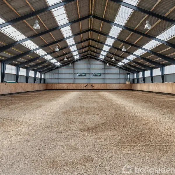 Ridehal med lysgråt sand underlag strækker sig under et stort metaltag med ovenlys. Væggene er af træ, omgivet af stålbjælker, og elektriske lamper hænger fra loftet.