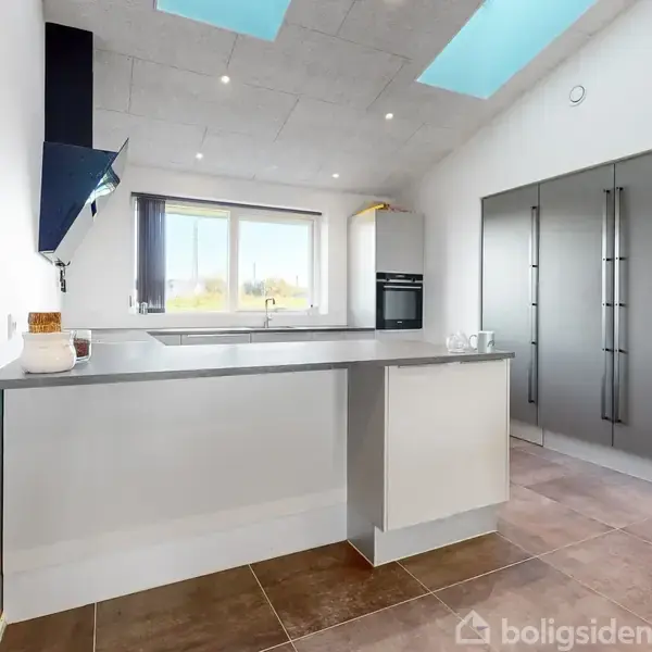 Et moderne køkken med en grå køkkenø og indbyggede skabe; sollys strømmer ind gennem et stort vindue. Loftet har store ovenlyspaneler, og gulvet er flisebelagt.