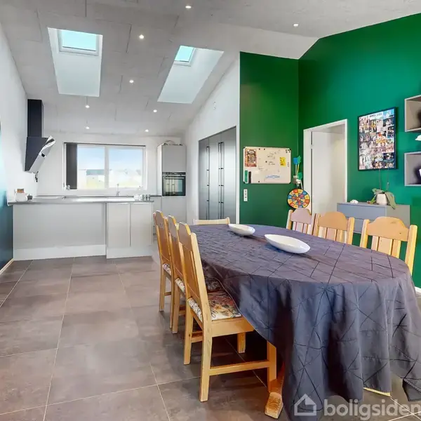 Lang spisebord med stole står centralt i moderne rum; køkken i baggrunden med grå elementer. Grøn væg til højre dekoreret med hylder og billeder.