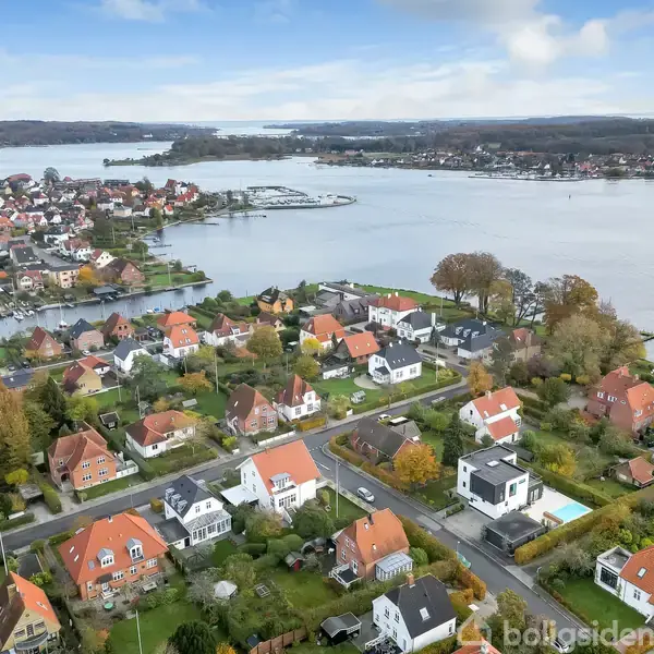Huse med røde tage står stille i et villakvarter ved vandet, omgivet af træer og grønne områder under en lys himmel.