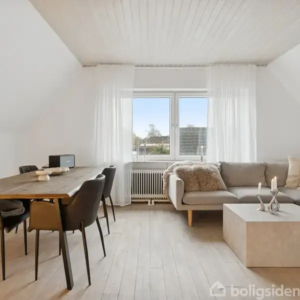 En stue med en stor, grå sofa og et træspisebord med flere sorte stole. Et stort vindue lader naturligt lys komme ind, omgivet af hvide gardiner.