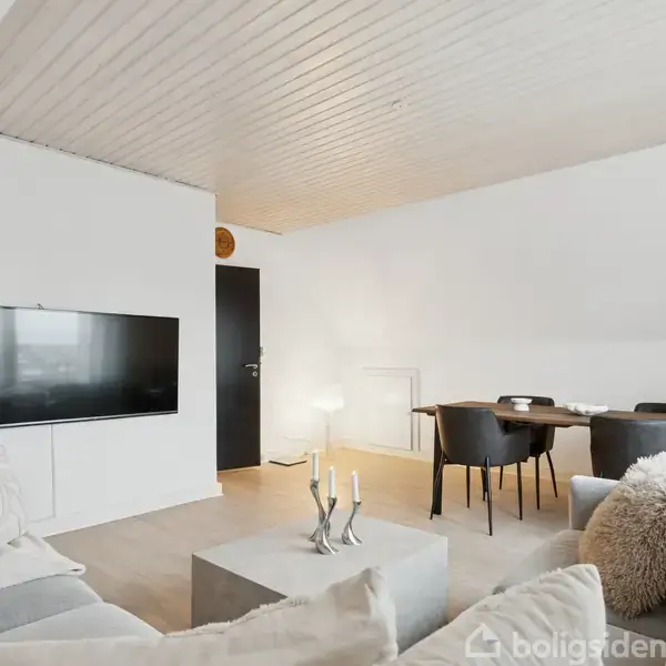 En stue med lyse vægge indeholder en grå sofa, rektangulært sofabord og vægmonteret fladskærms-TV. Et spisebord med stole står i baggrunden. En dør fører til et andet rum.