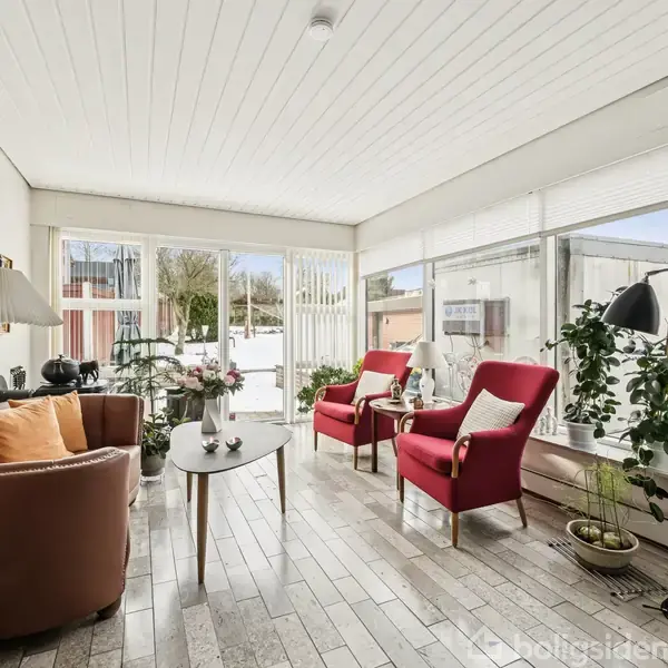 En stue med røde lænestole og en brun sofa omringer et bord. Store vinduer omgiver rummet, fyldt med planter og dekorationer, der giver udsigt til en have.
