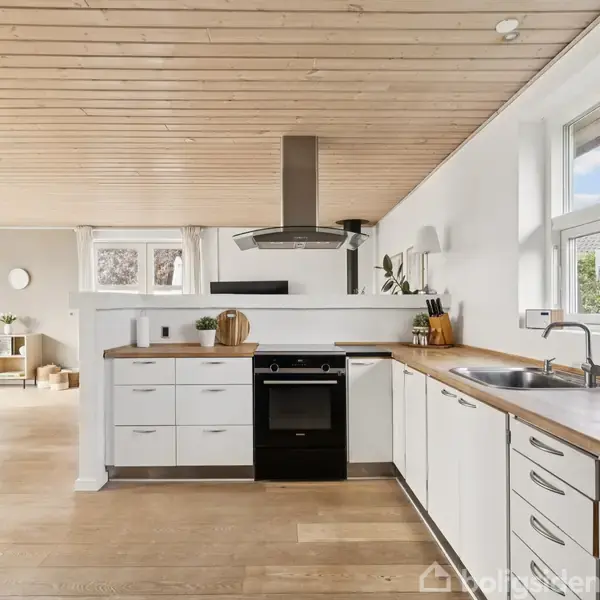 Et moderne køkken med hvide skabe, sort komfur og emhætte. Der er lysindfald fra vinduerne. Trægulve og lofter skaber en varm atmosfære. Alt udstråler funktionel enkelhed.