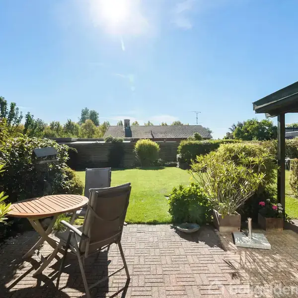 Et rundt træbord og en stol står stille på en solrig terrasse omgivet af grønne planter og en velplejet græsplæne bagved, mens himlen er klar og blå.