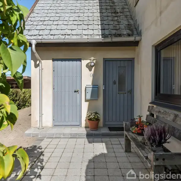 Husindgang med grå døre, en plante i en rød krukke ved siden af postkassen, en bænk med blomster foran et vindue. Belagt gårdsplads omkranset af grønne buske.