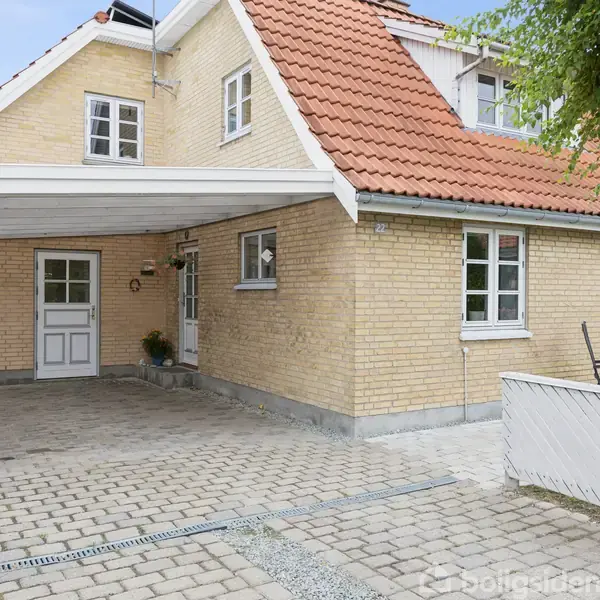 Hus med gule mursten og rødt gavltag. Carport til venstre og hvidt stakit til højre. Flisebelagt indkørsel foran. Grønne planter til højre.