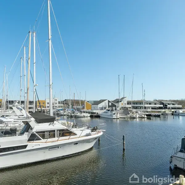 Både ligger stille ved en kaj i en marina, omgivet af moderne huse og klar blå himmel.